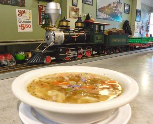 A bowl of soup sits on a table in front of a model train display in a cozy setting.