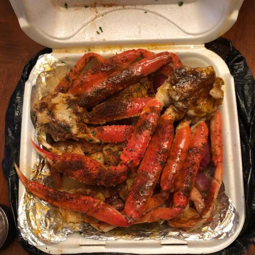 A takeout container filled with seasoned crab legs, potatoes, and spices, resting on a wooden table.