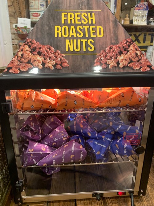 A display case for fresh roasted nuts, featuring colorful bags in orange and purple, with a sign on top.