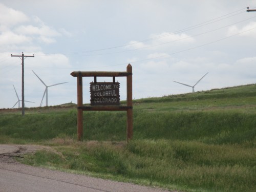 A wooden sign reading "Welcome to Colorful Colorado" stands beside a road, with wind turbines in the background.
