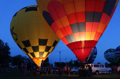 Two brightly lit hot air balloons at dusk, with people gathered around and other balloons in the background.