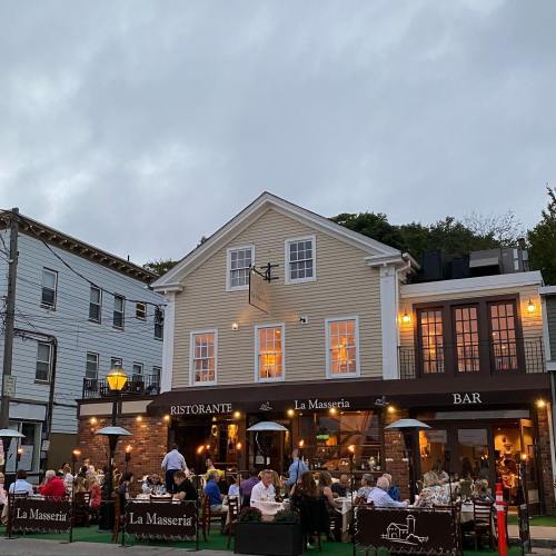 A cozy restaurant with outdoor seating, people dining, and a cloudy sky in the background. "La Masseria" is visible.