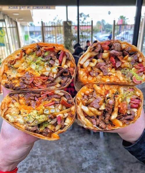Two large burritos filled with meat, cheese, potatoes, and vegetables, held up in front of a restaurant.