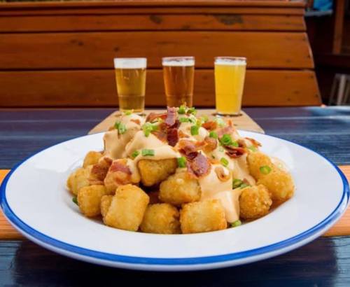 A plate of loaded tater tots topped with cheese, bacon, and green onions, with three glasses of beer in the background.