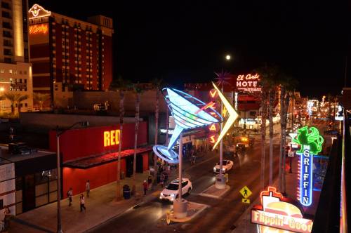 Vibrant neon signs illuminate a bustling street at night, with palm trees and a full moon in the background.