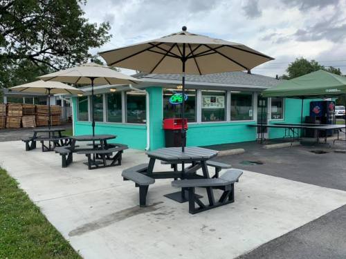 A turquoise food stand with outdoor seating and umbrellas, surrounded by a paved area and trees in the background.