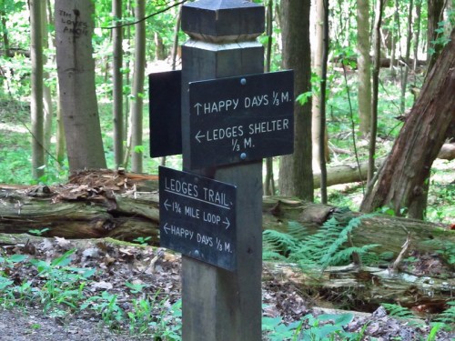 Trail signpost in a forest, indicating directions to Happy Days and Ledges Shelter, with distances marked.