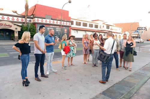 A diverse group of people gathers on a city street, listening to a speaker in front of shops and buildings.