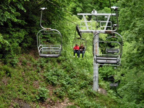 Two people sitting on a chairlift surrounded by lush green trees.