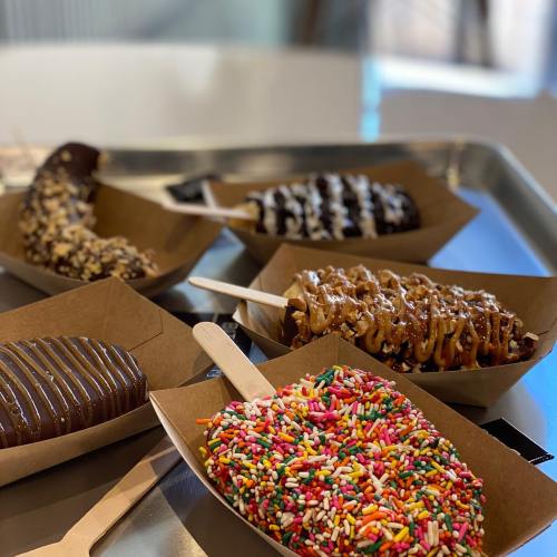 A variety of chocolate-covered bananas on wooden sticks, topped with sprinkles, nuts, and caramel, served in paper trays.