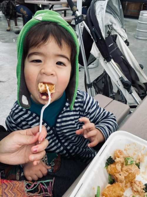 A young child wearing a green hat eagerly takes a bite of food from a fork held by an adult.