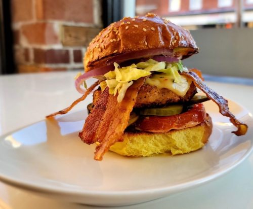 A delicious burger with bacon, lettuce, tomato, pickles, and cheese on a sesame seed bun, served on a white plate.
