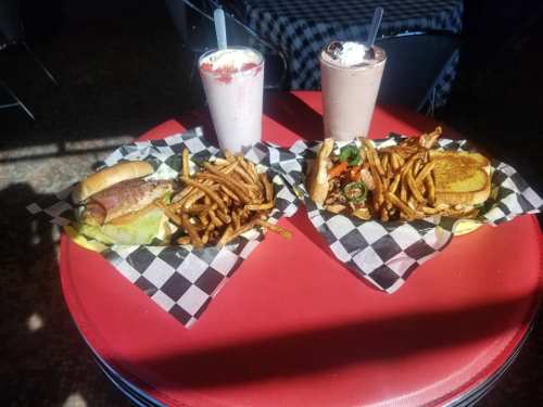 Two plates of food on a red table: a hot dog, fries, and a burger with a milkshake in the background.