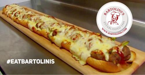 A long sub sandwich topped with melted cheese, peppers, and sausage, served on a wooden platter with a logo in the corner.