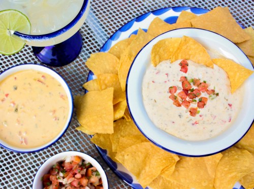 A colorful spread of tortilla chips with two bowls of queso and salsa, alongside a margarita with a lime wedge.