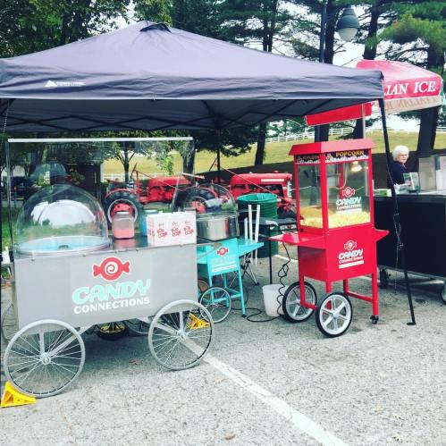 A colorful candy cart and popcorn stand under a tent at an outdoor event, surrounded by trees and parked vehicles.