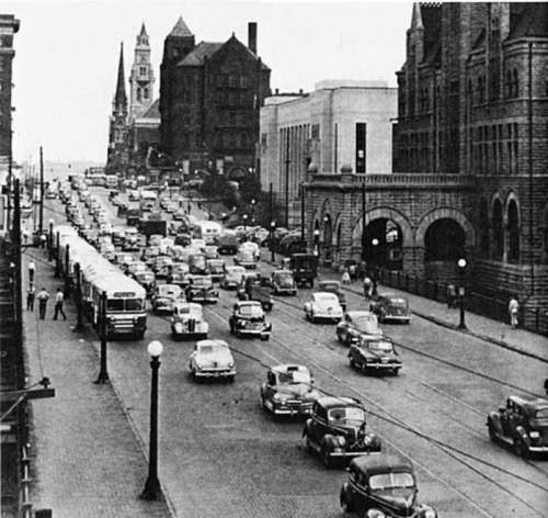 Historic black-and-white photo of a busy street with cars, buses, and pedestrians, featuring buildings and lampposts.