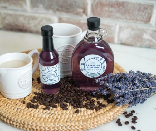 A cozy arrangement featuring lavender syrup, a small bottle, a mug, cream, coffee beans, and dried lavender on a woven mat.