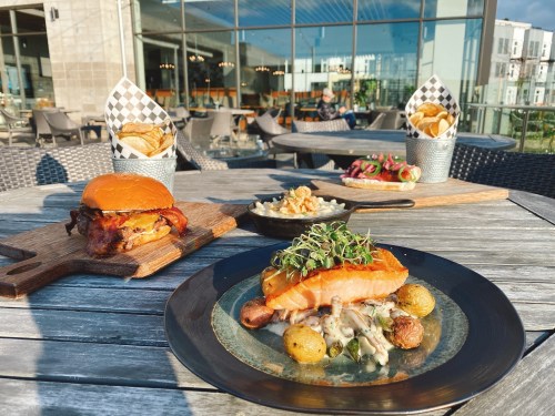 A beautifully arranged outdoor dining table featuring a salmon dish, a burger, mac and cheese, and two servings of fries.
