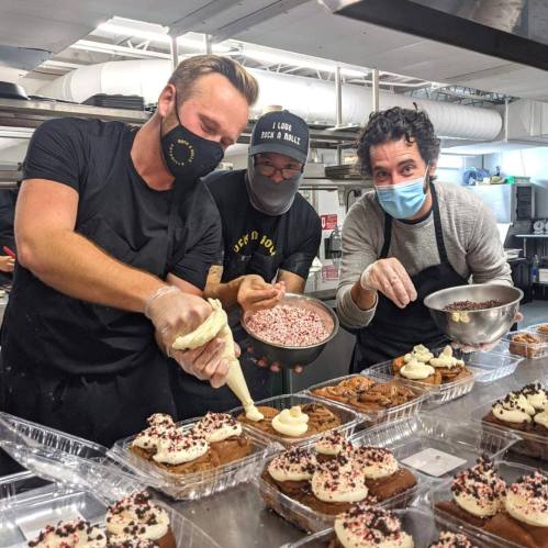 Three bakers decorate cupcakes in a kitchen, using piping bags and bowls of toppings, all wearing masks and aprons.