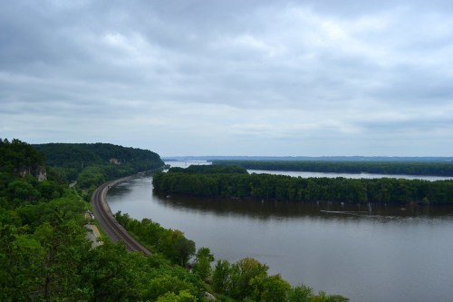 A winding river surrounded by lush greenery under a cloudy sky, with a train track along the riverbank.
