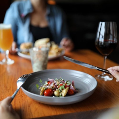 A close-up of a plated salad with cherry tomatoes and herbs, with drinks and another dish in the background.