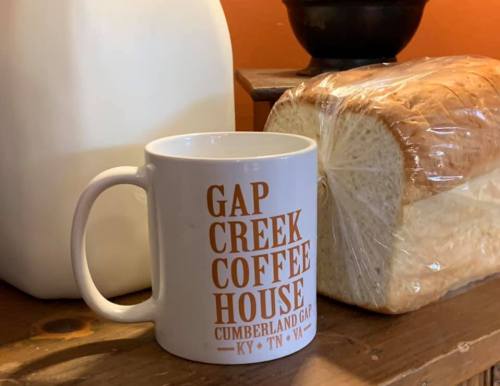 A white coffee mug with "Gap Creek Coffee House" text next to a jug of milk and a loaf of bread on a wooden surface.