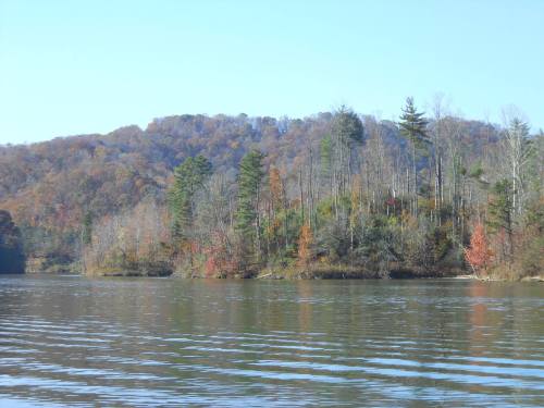 A serene lake scene with colorful autumn trees reflecting on the water, surrounded by rolling hills under a clear blue sky.