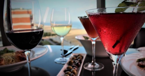 A table setting with various drinks, including red wine and cocktails, overlooking a beach view.