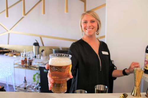 A smiling bartender holds a glass of beer at a stylish bar with gold accents in the background.