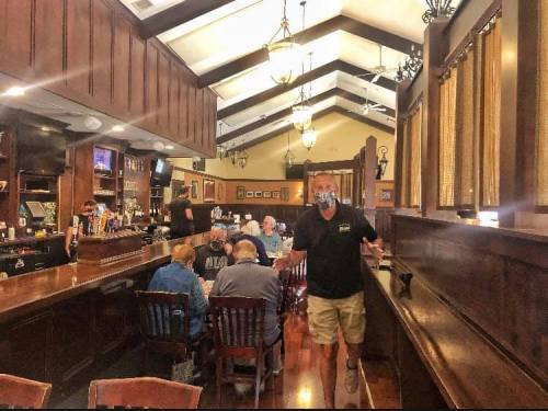 A lively bar scene with patrons seated at tables and a masked server interacting with guests in a cozy setting.