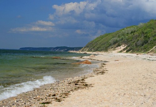 A serene beach scene with gentle waves, rocky shore, and lush green hills under a cloudy sky.