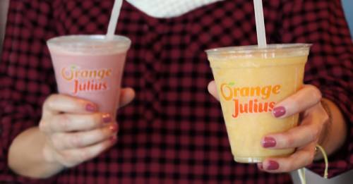 A person holding two cups of Orange Julius drinks, one pink and one yellow, with a checkered shirt in the background.