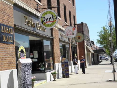 A street view of shops with signs for "Perks Corner" and "The Pretty Pickle," featuring brick buildings and pedestrians.