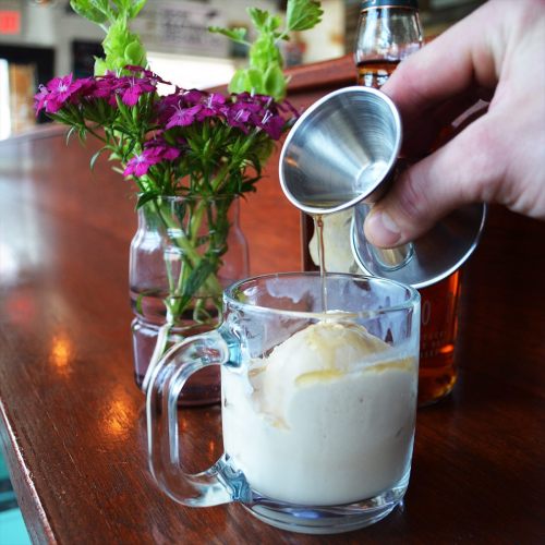 A hand pours syrup over a glass of ice cream, with a small vase of flowers in the background.