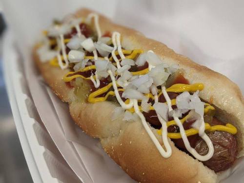 A close-up of a hot dog in a bun, topped with mustard, mayonnaise, onions, and pickles, served in a white container.