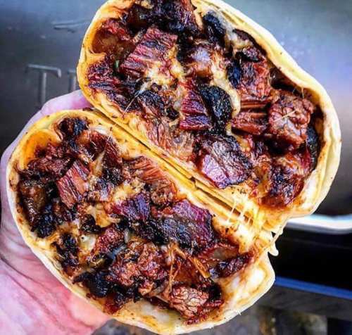 A hand holding a halved burrito filled with shredded beef and melted cheese, showcasing a smoky, grilled texture.