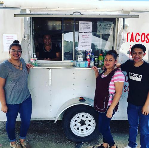 A food truck with a smiling vendor and three customers posing in front, enjoying their time together.
