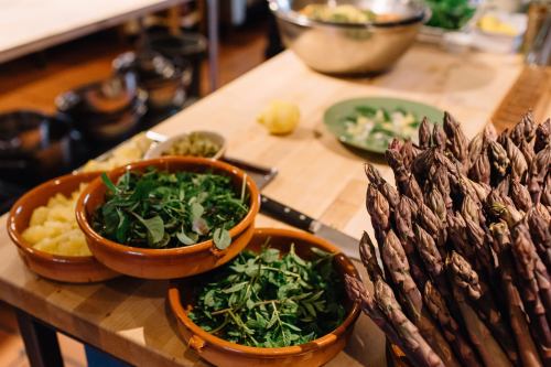 Fresh herbs and asparagus on a wooden kitchen counter, with bowls of greens and a bowl of lemon in the background.