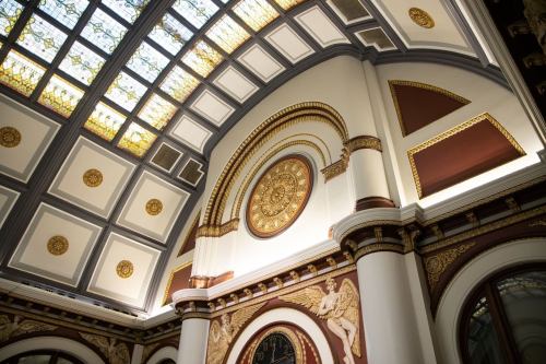 Interior of a grand building featuring ornate architecture, a decorative clock, and a colorful stained glass ceiling.