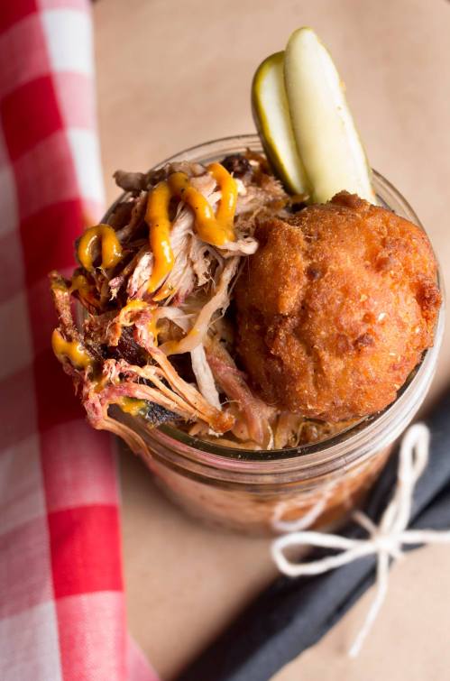 A jar filled with pulled pork, a cornbread ball, pickles, and drizzled mustard, set on a checkered tablecloth.