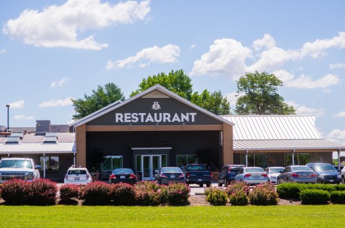 A restaurant building with a large sign, surrounded by green grass and parked cars under a blue sky with clouds.