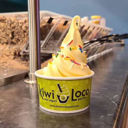 A swirl of yellow frozen yogurt topped with colorful sprinkles in a green Kiwi Loco cup.