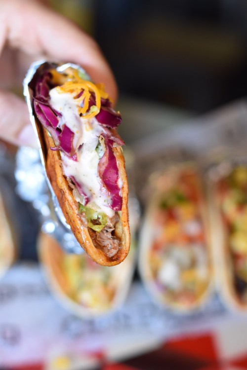 A close-up of a hand holding a taco filled with meat, lettuce, cheese, and sauce, with more tacos blurred in the background.