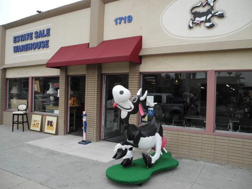 A whimsical black and white cow statue outside an estate sale warehouse with a red awning and large windows.