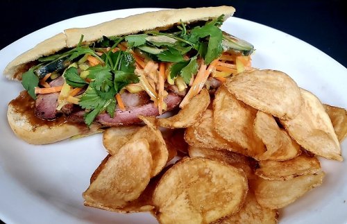 A sandwich filled with sliced meat, fresh herbs, and vegetables, served with a side of crispy potato chips.