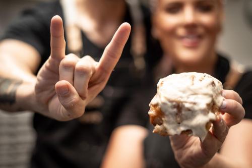 A person holds up a cinnamon roll while another makes a rock sign gesture in the background, both smiling.