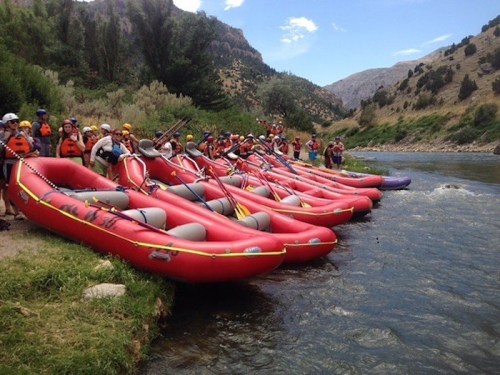A line of red rafts on a riverbank, with people in life jackets preparing for a rafting adventure in a scenic landscape.