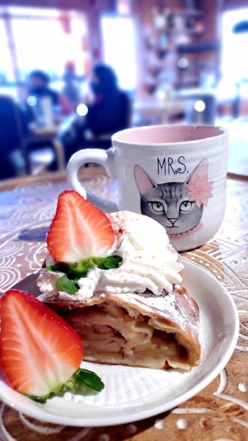 A slice of apple strudel topped with whipped cream and strawberries, next to a cat-themed mug labeled "MRS." in a cozy café.