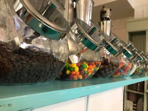 A row of glass jars filled with various colorful candies and chocolate chips on a turquoise shelf.